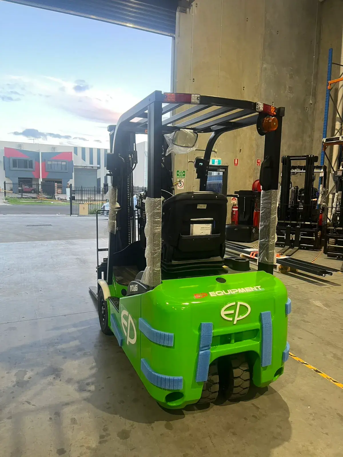 Rear three-quarter view of an EP Equipment TCL152 1.5T three-wheel electric forklift at a warehouse roller door, showing the overhead guard, rear work lights and beacon